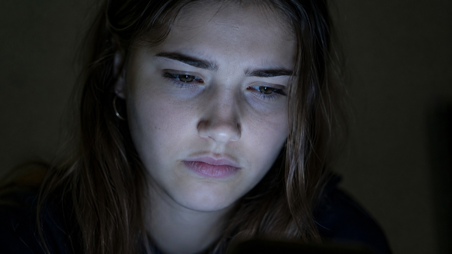 A young woman's face lit by a phone screen at night, her expression skeptical rather than alarmed.
